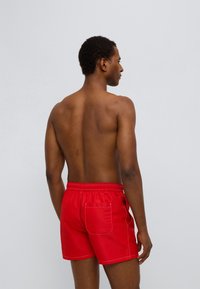 Κόκκινα μαγιό σορτς με ελαστική μέση και τσέπη στο πίσω μέρος, με αντίθετη ραφή. Λείο, ελαφρύ υλικό. Το μοντέλο εμφανίζεται από πίσω.