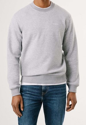 Man draagt een lichtgrijze trui met ronde hals over een wit shirt, gecombineerd met een blauwe spijkerbroek, en staat met ontspannen handen langs zijn zij.