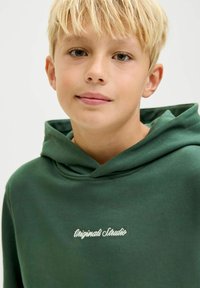 Zelena dukserica od mekog materijala, s velikom kapuljačom i izvezenim logom "Originals Studio" u bijeloj boji na prsima.