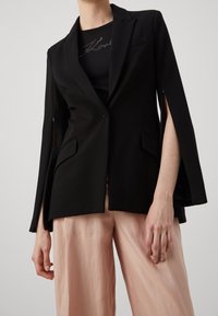 Blazer negro con mangas abiertas, corte entallado, cierre de un botón y dos bolsillos delanteros, combinado con pantalones anchos de pierna en rosa claro.