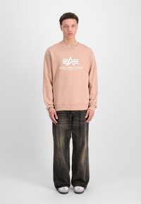 Sudadera rosa claro con el logo de "Alpha Industries", combinada con jeans de pierna ancha en gris oscuro y zapatillas deportivas blancas.