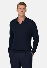 Homme aux cheveux blonds courts, portant un pull à manches longues côtelé bleu marine foncé et un pantalon assorti, debout les mains dans les poches.