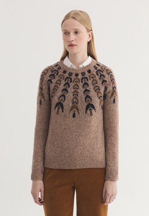 Femme portant un pull en laine marron avec un motif symétrique noir et marron foncé, un col de chemise blanc et un pantalon marron sur fond blanc.