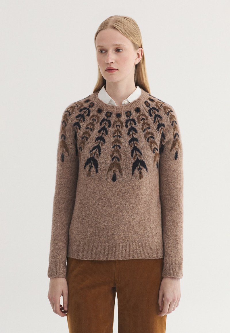 Femme portant un pull en laine marron avec un motif symétrique noir et marron foncé, un col de chemise blanc et un pantalon marron sur fond blanc.
