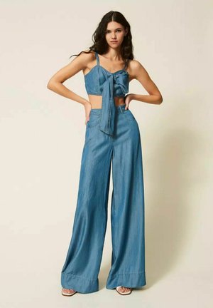 Giovane donna con pantaloni larghi in denim blu e top corto abbinato con grande fiocco sul davanti, in piedi con le mani sui fianchi davanti a uno sfondo semplice.