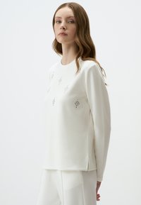 Wit langemouwig top van zacht materiaal, met decoratieve rhinestones in een bloemmotief op de voorkant en zijsplitten voor gemakkelijke draagbaarheid.
