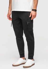 Personne portant un pantalon cargo jogger noir avec des poches zippées, des baskets blanches et un t-shirt blanc, montré de la taille vers le bas.