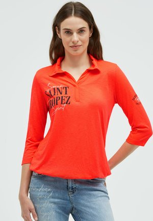 Woman with long brown hair wearing a bright orange three-quarter sleeve shirt with "Saint Tropez" text and blue jeans with script writing.