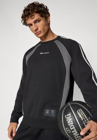 Sudadera negra con detalles en gris, cuello redondo y puños acanalados, con el logo de Champion en el pecho y una etiqueta en el dobladillo.