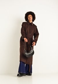 Cappotto marrone con bottoni doppi, indossato su pantaloni larghi blu scuro. Modella che tiene una borsa nera a forma curvata, in piedi su uno sfondo bianco.