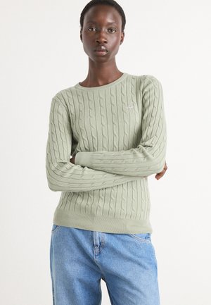 Lichtgroene kabelgebreide sweater met lange mouwen en geribde manchetten, met een klein logodetail op de borst. Gepaard met een blauwe spijkerbroek.