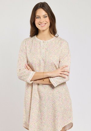 Femme souriante aux cheveux bruns portant une robe légère à motif floral avec des manches trois-quarts, debout les bras croisés devant un fond uni.