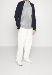 Granatowy sweter z dzianiny, jasnoszara koszula w paski z guzikami, białe spodnie o szerokich nogawkach i białe tenisówki. Swobodna stylizacja z warstwami w kontrastowych kolorach.