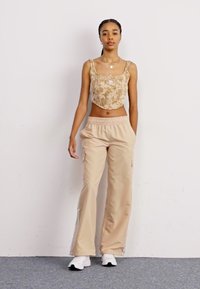 Μπεζ λουλουδάτο αμπιγιέ μπλουζάκι με λεπτές τιράντες και ταιριαστές μπεζ φαρδιές παντούφλες. Μοντέλο που φοράει λευκά αθλητικά παπούτσια, στέκεται μπροστά σε απλό φόντο.