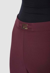 Pantaloni bordeaux con una texture liscia, un design aderente e una fibbia con logo dorata in vita. Sagoma su misura e lineare.