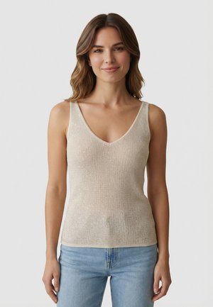 Femme aux cheveux bruns mi-longs portant un haut en tricot beige sans manches et un jean bleu clair, souriant devant un fond uni.