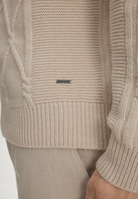 Beige gestrickter Pullover mit Kabelmustern und geripptem Saum. Verfügt über ein kleines Metallschildchen. Glatte Textur und lockere Passform.