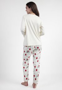 Mujer de pie descalza, vistiendo una blusa larga color marfil y pantalones de pijama blancos con personajes de cómic en rojo y negro.