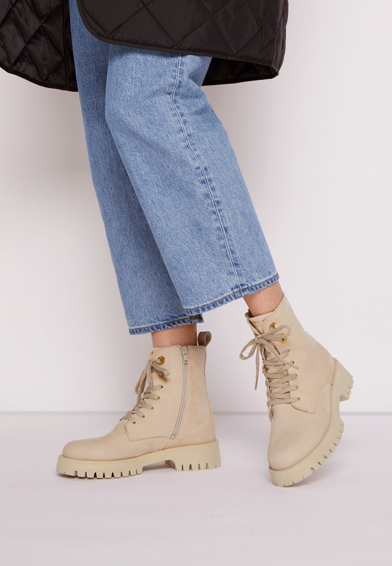Bottes beiges à la cheville avec lacets, fermeture éclair latérale et semelle épaisse. Fabriquées en daim avec une texture lisse et une boucle à l'arrière pour un enfilage facile.