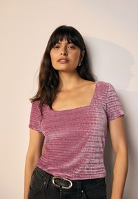 Getailleerd kortermig topje van paars, gestructureerd stof met een vierkante halslijn. Gecombineerd met zwarte broek en een riem met een ronde gesp.