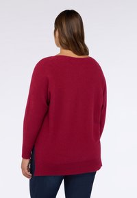 Roter Pullover mit lockerem Sitz, rundem Ausschnitt und gerippten Bündchen. Seitliche Schlitze am Saum. Weiche Textur. Kombiniert mit dunkelblauer Jeans.