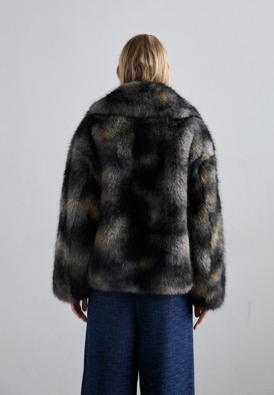 Osoba z blond włosami, ubrana w ciemną, melanżową kurtkę futrzaną i niebieskie, teksturowane spodnie o szerokich nogawkach, stojąca na jednolitym, jasnym tle.