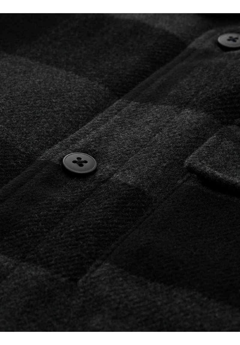 Nahaufnahme einer schwarz und dunkelgrau karierten Wolljacke mit zwei schwarzen Knöpfen und einer kleinen Klappentasche.