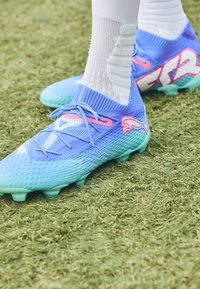 Blue soccer cleats with gradient design, featuring a textured mesh upper, tonal laces, and green studded outsole. Notable pink accents.