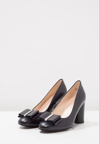 Scarpe con tacco alto in pelle nera, con punta arrotondata, dotate di dettagli in fiocco e particolari metallici sul davanti. Texture liscia e tacco robusto.
