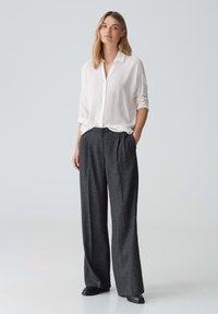 Camicia bianca con maniche arrotolate, abbinata a pantaloni a vita alta grigi scuri con pieghe e calzature nere. Design semplice e sartoriale.