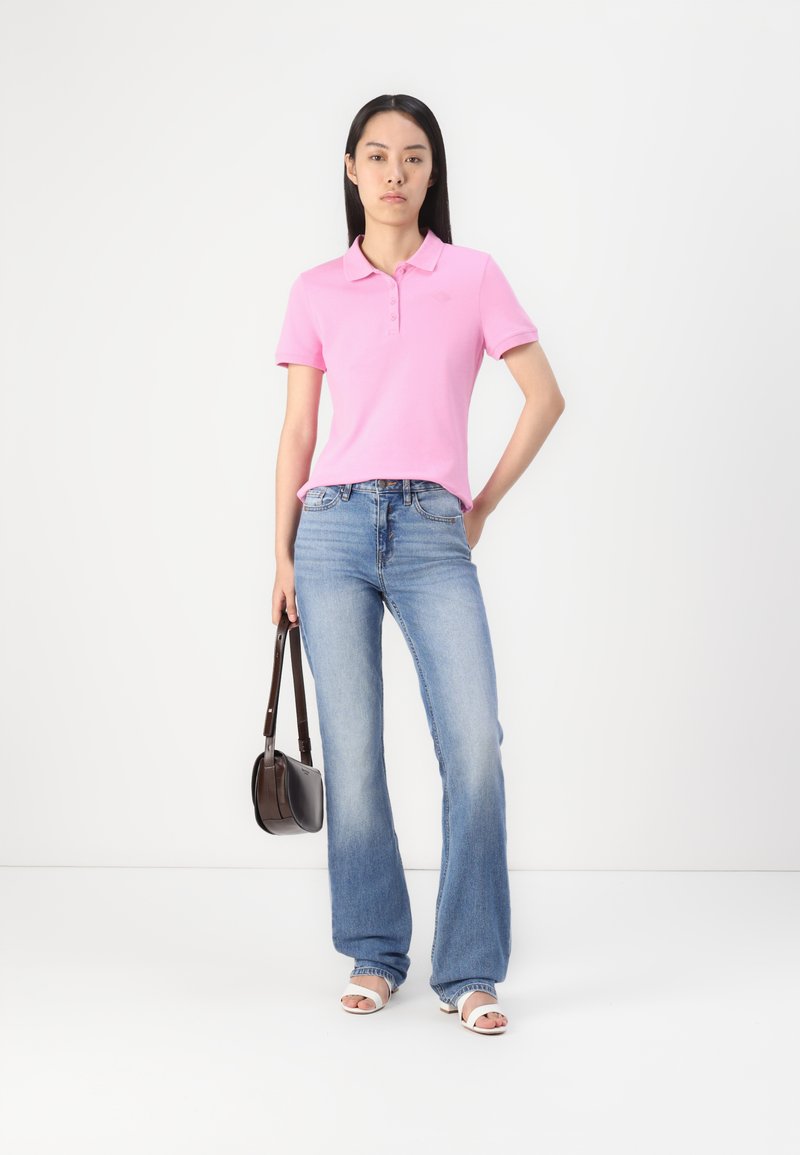 Rosa Polohemd mit Kragen und drei Knöpfen, kombiniert mit hellblauen Schlagjeans. Das Model hält eine braune Handtasche und trägt weiße Sandalen.