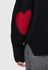 Czarny sweter dzianinowy z długimi rękawami, z dużym, puszystym czerwonym sercem na lewym rękawie. Teksturowana tkanina, luźny krój.