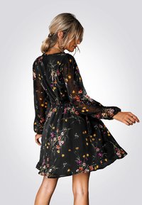 Zwarte bloemenjurk met lange doorschijnende mouwen, een gathered taille en kleurrijke bloemenpatronen in geel, roze en wit.