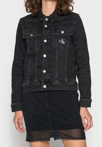 Svart jeansjacka med silverfärgad hårdvara, innehållande framfickor, knappstängning och en Calvin Klein-logotyp på vänster bröst.