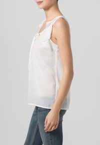 Blusa bianca senza maniche con un nodo al colletto, caratterizzata da dettagli arricciati sulle spalle, realizzata in tessuto leggero e trasparente.