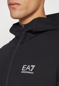 Musta vetoketjullinen huppari korkealla kauluksella, jossa on valkoinen EA7 Emporio Armani -logo rinnassa ja sileä kangaspinta.