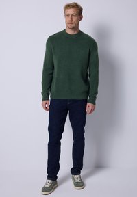 Suéter de punto verde oscuro con cuello redondo acanalado y puños, combinado con jeans oscuros y zapatillas verdes, de pie frente a un fondo blanco.