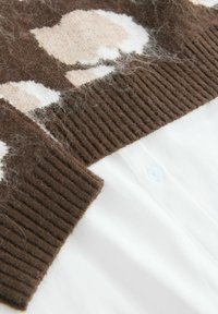 Brązowy wełniany sweter z dzianiny z jasnym wzorem, z ribbowym wykończeniem i widoczną białą koszulą pod spodem, z detalem guzika.