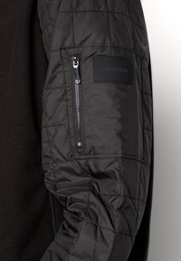 Zwarte gewatteerde jas met een gladde stof op het lichaam, voorzien van een ritszak op de linkermouw en een Calvin Klein logo patch.