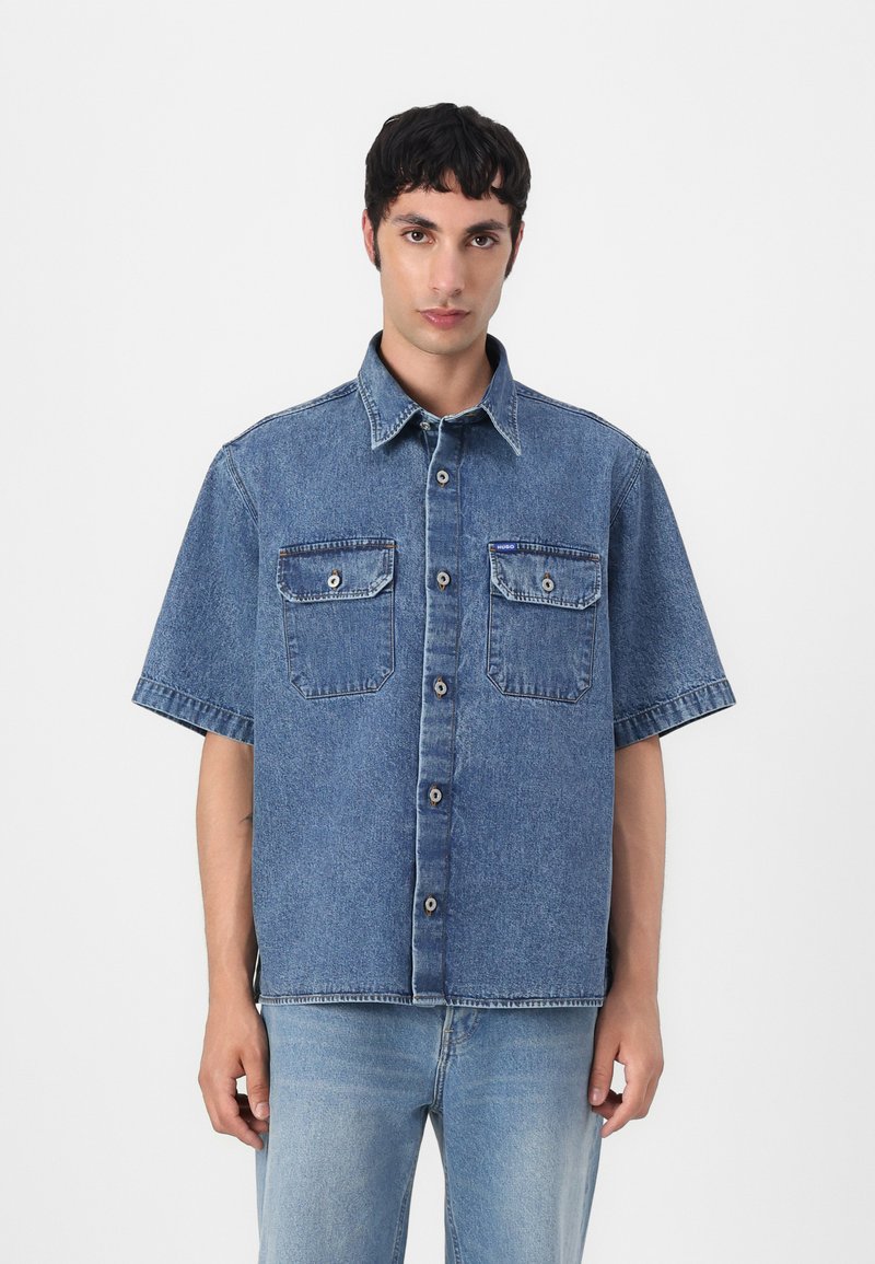Világoskék denim rövid ujjú ing, gombos elülső résszel, két mellzsebbel és laza szabással. Simácska felület, látható varrásokkal.