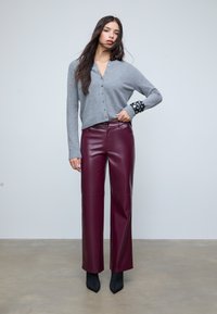 Vrouw in een grijze knopen cardigan, bordeaux leren broek, zwarte enkellaarsjes en een gespannen zwarte polsband, staande voor een neutrale achtergrond.