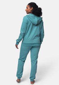 Felpa con cappuccio color teal e pantaloni jogger abbinati in morbido tessuto. La felpa presenta un cappuccio, polsini a costine e una vestibilità rilassata.