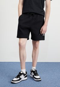 Černé bavlněné šortky s páskem na stažení, bočními kapsami a ohrnutými lemy. Kombinovány s černobílými teniskami a bílými ponožkami.