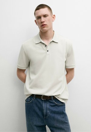 Jonge man met kort haar, gekleed in een lichtbeige poloshirt en blauwe jeans, staand tegen een effen witte achtergrond.