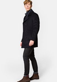 Czarny wełniany płaszcz typu peacoat z podwójnymi guzikami, szary kołnierz, w połączeniu z ciemnymi zniszczonymi dżinsami i brązowymi botkami Chelsea.