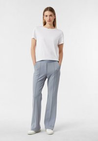 Biała bawełniana koszulka zestawiona z jasnoszarymi, dopasowanymi spodniami z plisami i paskiem z logo. Model stoi z rękami w kieszeniach.