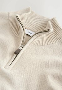 Sweatshirt zippé beige clair en tissu doux, avec un col et des poignets côtelés. Fermeture éclair agrémentée d'une tirette marron. Étiquette de la marque visible.