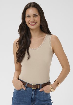 Femme souriante aux longs cheveux noirs portant un débardeur beige orné de dentelle, un jean bleu, une ceinture marron et des bracelets en or, sur un fond uni.