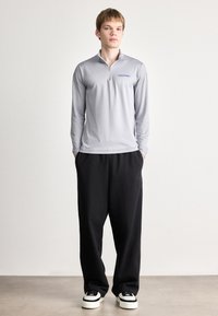 Γκρι μακρυμάνικη μπλούζα με φερμουάρ ως το ένα τέταρτο, που διαθέτει μπλε λογότυπο. Μαύρα φαρδιά παντελόνια, ολοκληρωμένα με αθλητικά παπούτσια με λευκή σόλα.