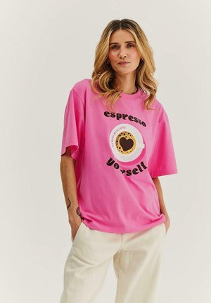 Kobieta z falującymi blond włosami, ubrana w luźny różowy t-shirt z grafiką filiżanki kawy i tekstem "espresso yourself", w połączeniu z jasnymi spodniami.